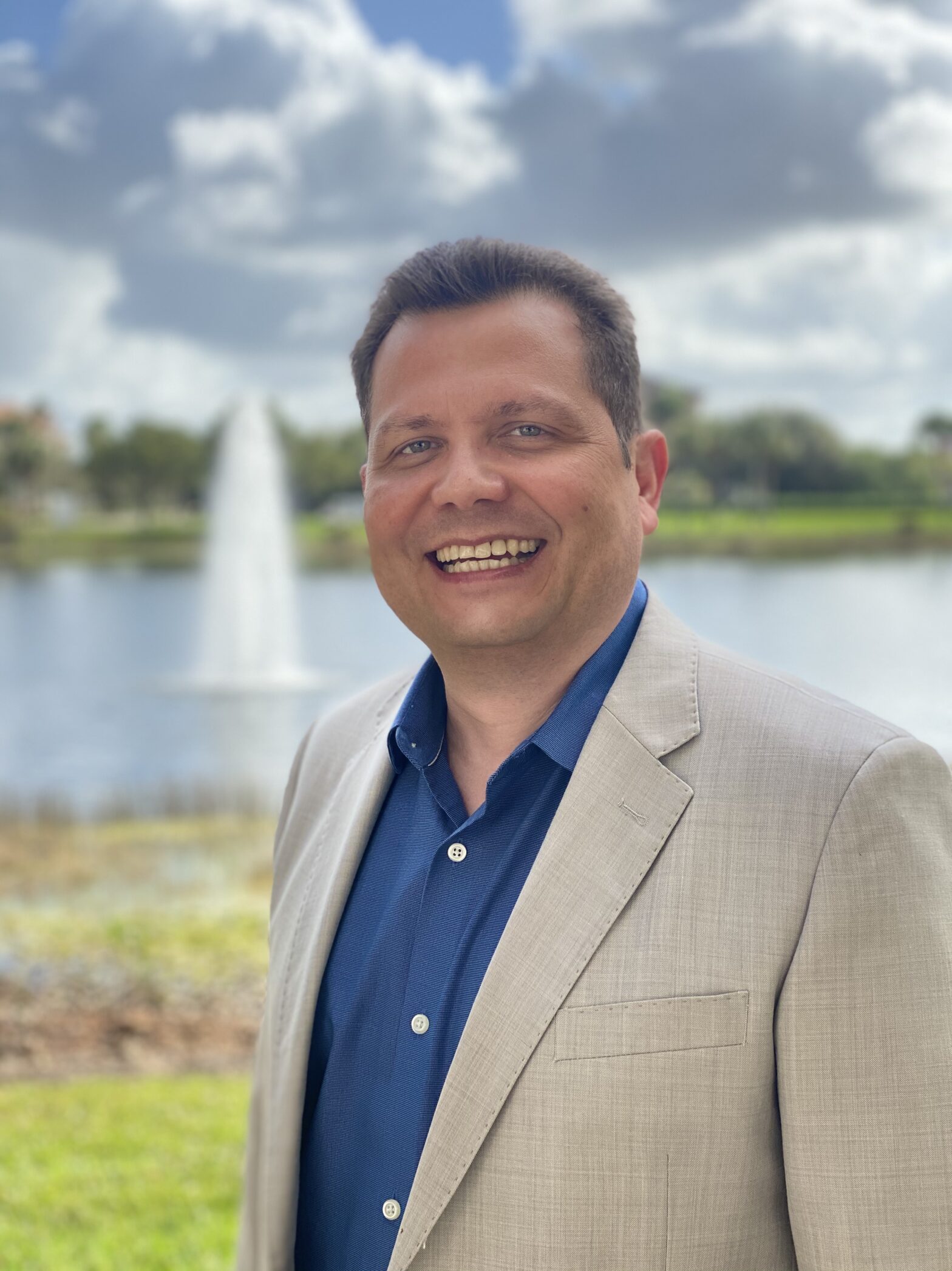 Smiling man in a beige blazer by a lake with a fountain.